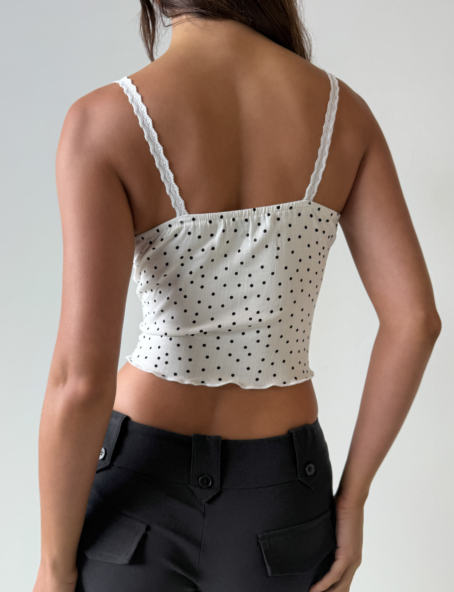 A woman is shown from the back wearing the Third Party BAILEE CAMI TOP - WHITE POLKA with lace trim, paired with black high-waisted pants against a plain light background.
