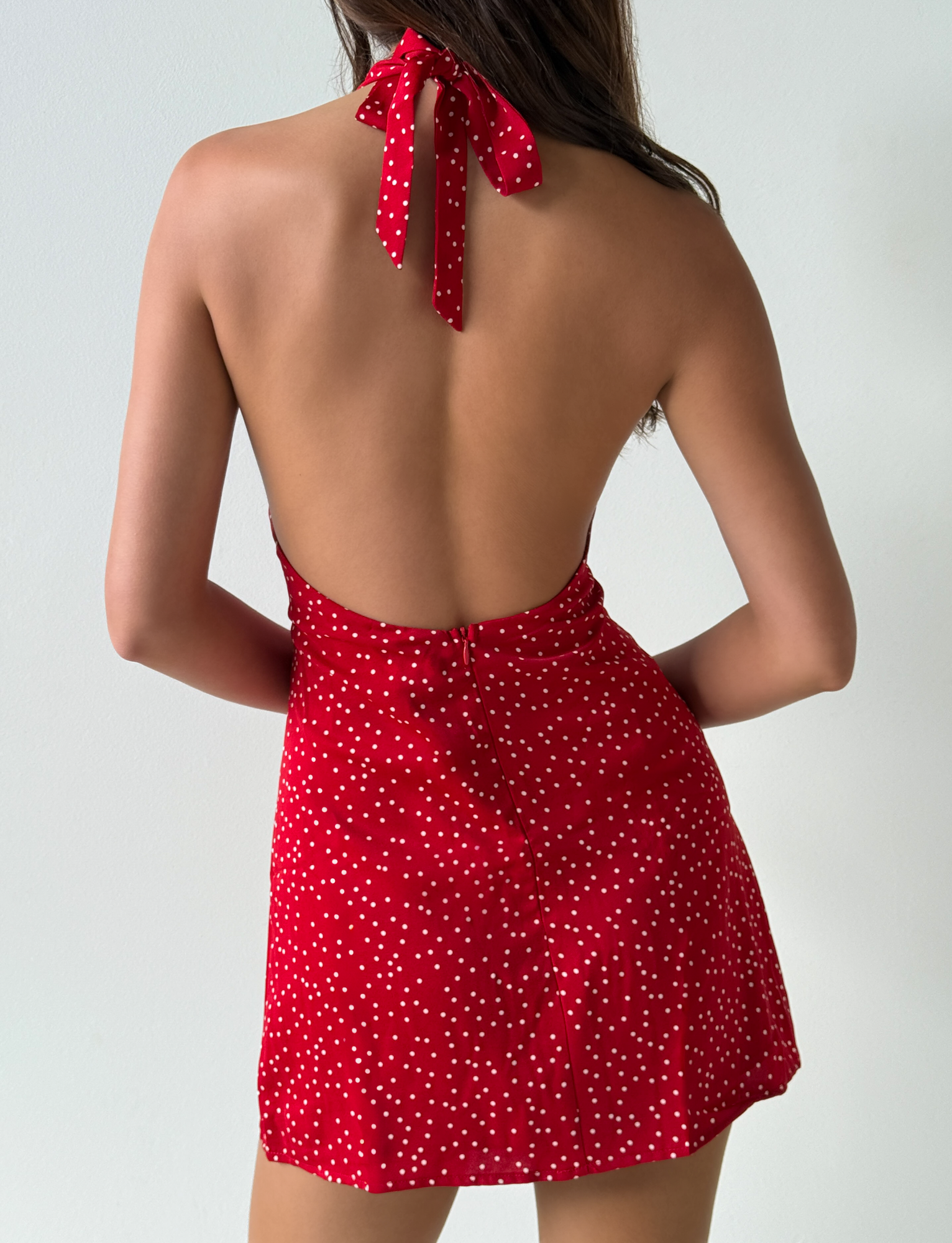 A woman with long brown hair, facing away, models the DYLAN HALTER MINI DRESS - RED POLKA by Third Party. The red dress features white polka dots and a halter neckline. She stands hands-on-hips against a simple, light backdrop—ideal for wholesale inspiration.