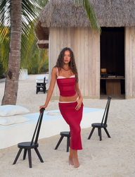 A woman models the TIGERMIST CALINE MIDI SKIRT - RED, paired with a matching tube top, barefoot on sandy ground among palm trees, black chairs, white cushions, and a thatched-roof wooden hut in the background.
