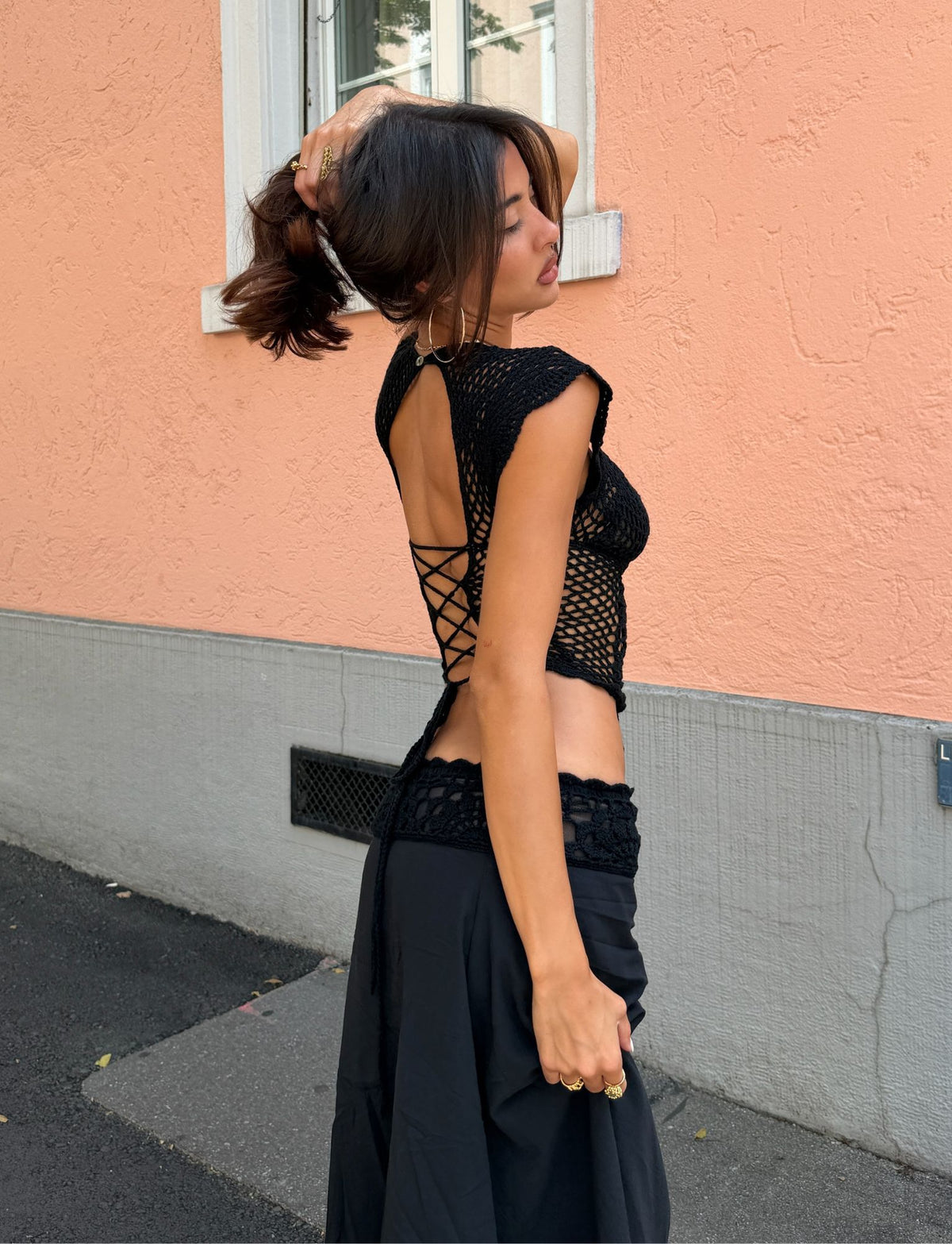 A woman stands outdoors against a peach-colored wall, embodying effortless resort style in the TIGERMIST SASHA CROCHET TOP - BLACK and a matching skirt. She lifts her hair with one hand and gazes to the side.