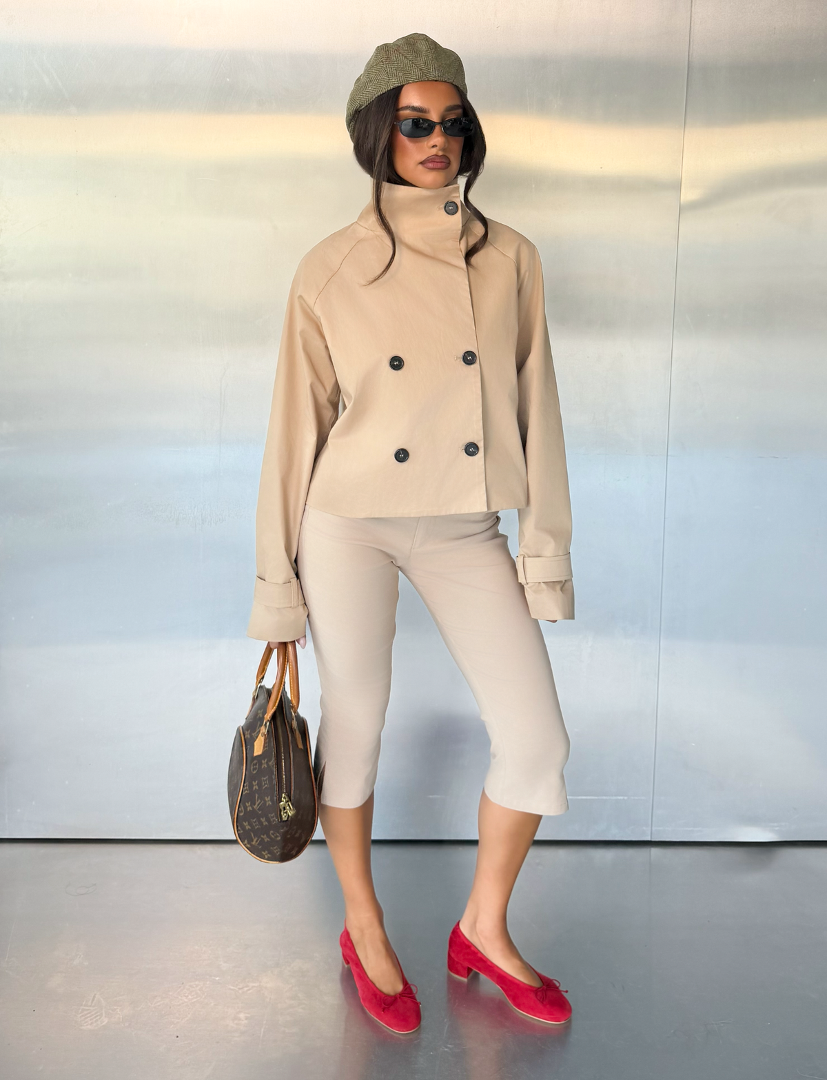 A woman wearing the HAYLEY CROPPED TRENCH JACKET in khaki by TIGERMIST, along with cropped beige pants, red shoes, a gray beret, sunglasses, and holding a brown handbag stands against a metallic wall.