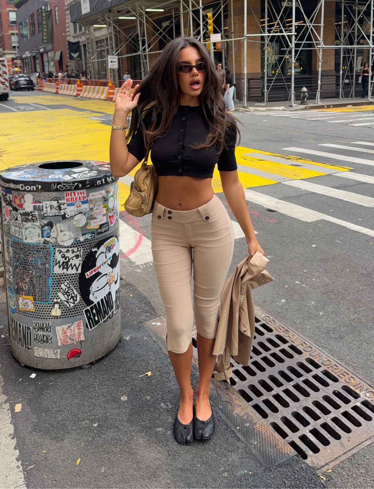 A woman with long hair stands on a city street, wearing sunglasses, a black crop top, and the TIGERMIST ARIA CAPRI - KHAKI, paired with black shoes. She holds a tan jacket and a gold purse. A graffiti-covered trash can and nearby construction are visible.