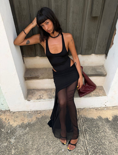 A woman with straight dark hair sits on a stone step wearing the TIGERMIST LILIANA-SUMMER MAXI DRESS in black, paired with black sandals and holding a burgundy bag, as she rests her head on one hand and gazes thoughtfully to the side.