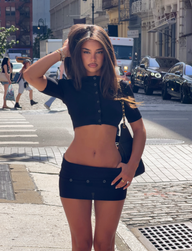 A woman in a black crop top and mini skirt stands confidently on a city street, layered with the ELSIE CARDIGAN - BLACK by TIGERMIST. The vibrant city backdrop of cars, people, and buildings highlights her effortlessly chic style.