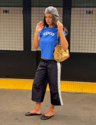A woman stands on a subway platform wearing a gray beret, a blue “BLESSED” T-shirt, and TIGERMIST’s SOPHIA WINDBREAKER CAPRI in navy and white. She completes her look with black flats and carries a tan purse. The background features tile walls and the number 23.