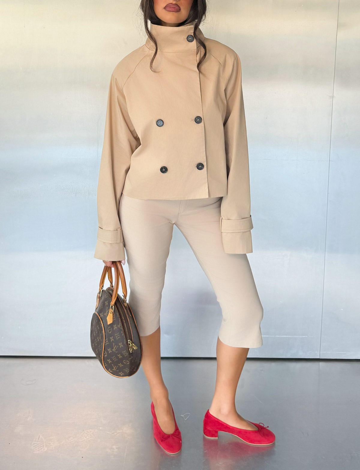 A woman wears the TIGERMIST HAYLEY CROPPED TRENCH JACKET in khaki with cropped pants, holding a brown handbag and matching bright red flat shoes, standing against a plain light-colored background.