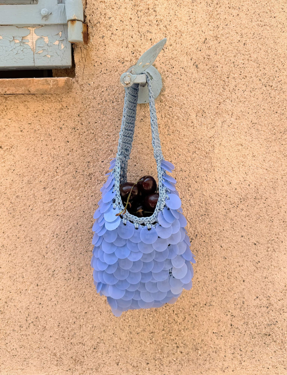 The CLEO BAG - BRIGHT BLUE by TIGERMIST, featuring circular crochet scales, hangs on a beige wall while holding a handful of dark cherries. Perfect as a chic beach bag, it sits just beneath a partially visible blue wooden shutter in the top left corner.