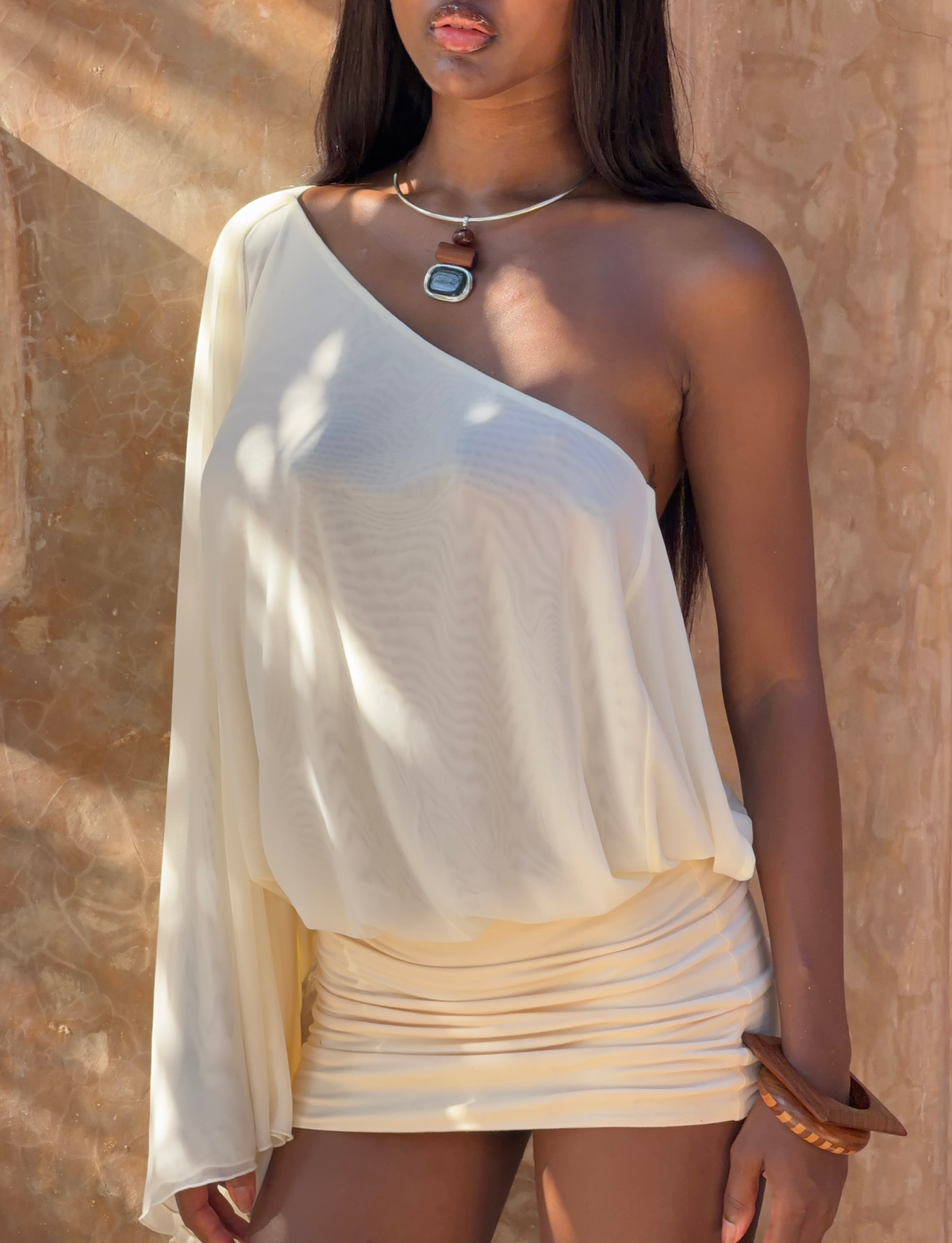 A woman in the TIGERMIST CAMILLE MINI DRESS - CREAM, styled with a necklace and bracelet, stands against a sunlit, textured wall.