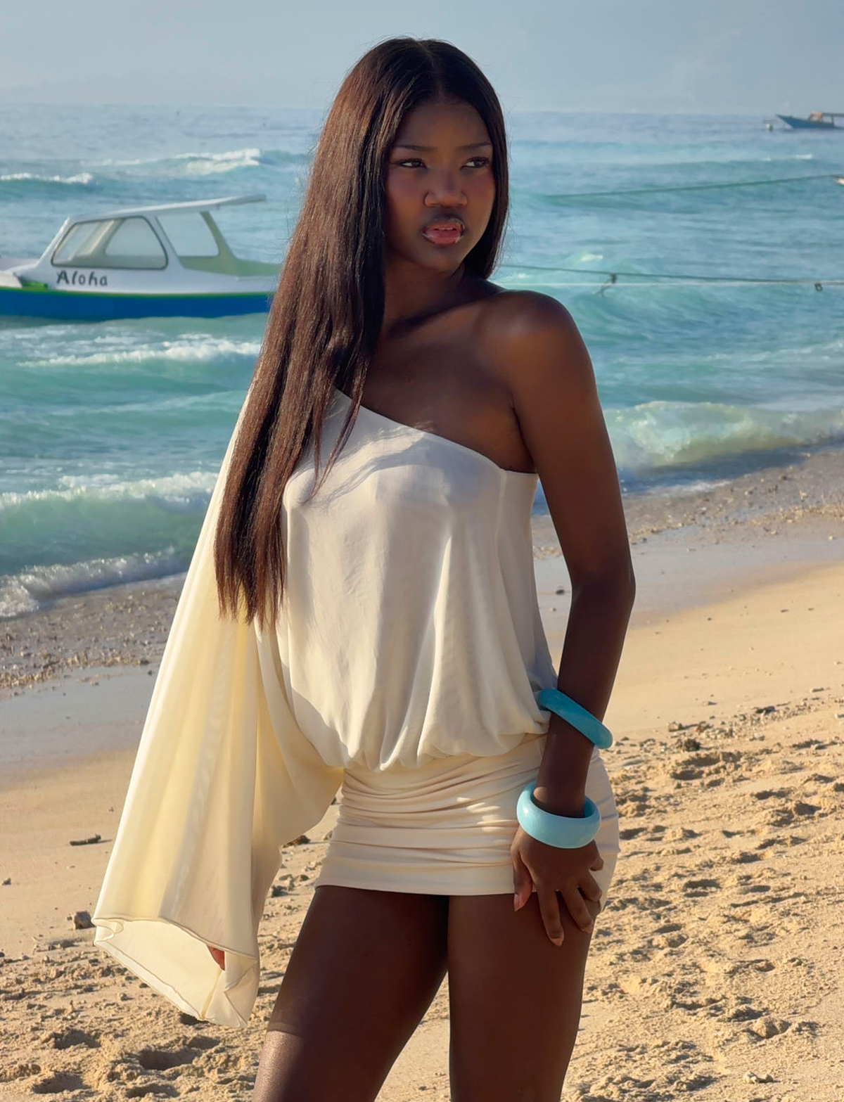 A woman in a cream one-shoulder dress wears the TIGERMIST MIKAH BANGLE SET - AQUA, standing on a sandy beach by clear water and a boat under the sunny sky.