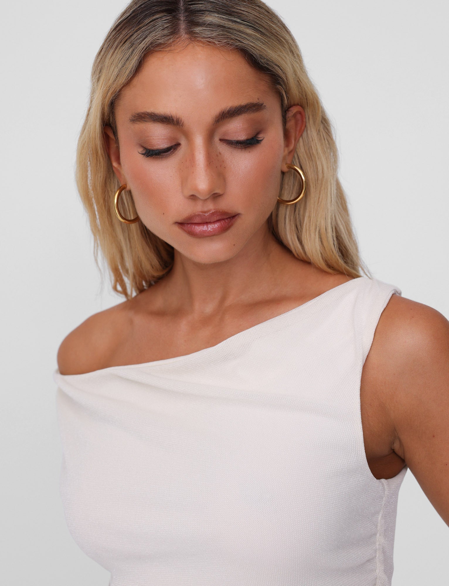A woman with long blonde hair gazes downward, adorned in a white one-shoulder top and the elegant PIPER EARRINGS - GOLD by TIGERMIST, lightweight and hypoallergenic hoops that stand out against the plain background.