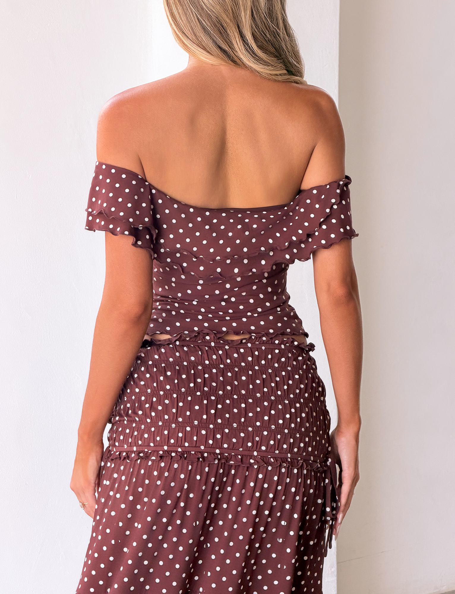 A woman with blonde hair faces away, wearing the TIGERMIST DENALI TOP - BROWN DOT. The off-shoulder top features polka dots, ruffled sleeves, and a fitted ruched waist against a plain, light background.