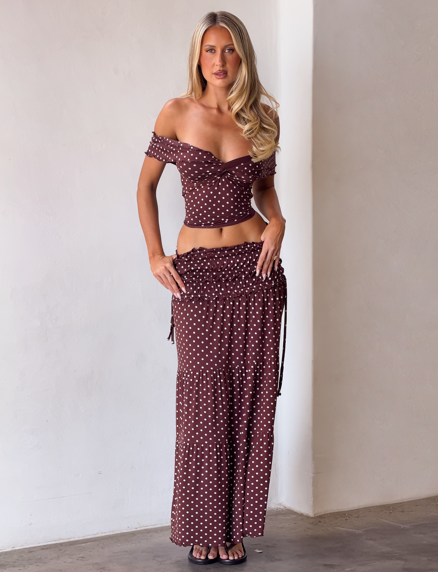 A woman stands against a white wall in the TIGERMIST DENALI TOP - BROWN DOT, featuring playful ruffled off-shoulder sleeves and a matching long skirt. She pairs the outfit with black open-toe heels and wears her long blonde hair loose.