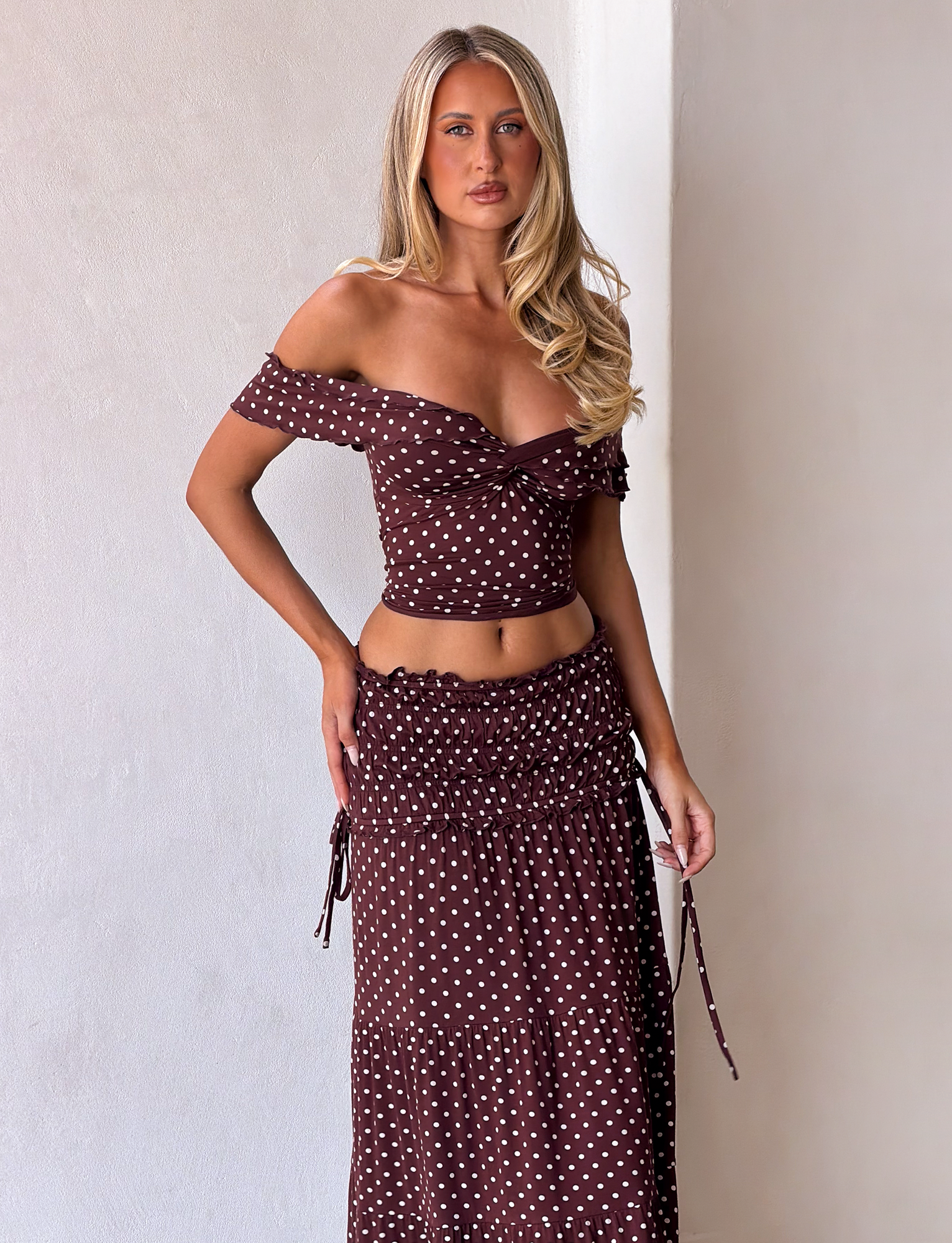 A woman with long, wavy blonde hair and a neutral expression wears a brown off-the-shoulder crop top and the TIGERMIST DENALI MAXI SKIRT - BROWN DOT, a soft modal polka dot maxi skirt, standing against a white wall.