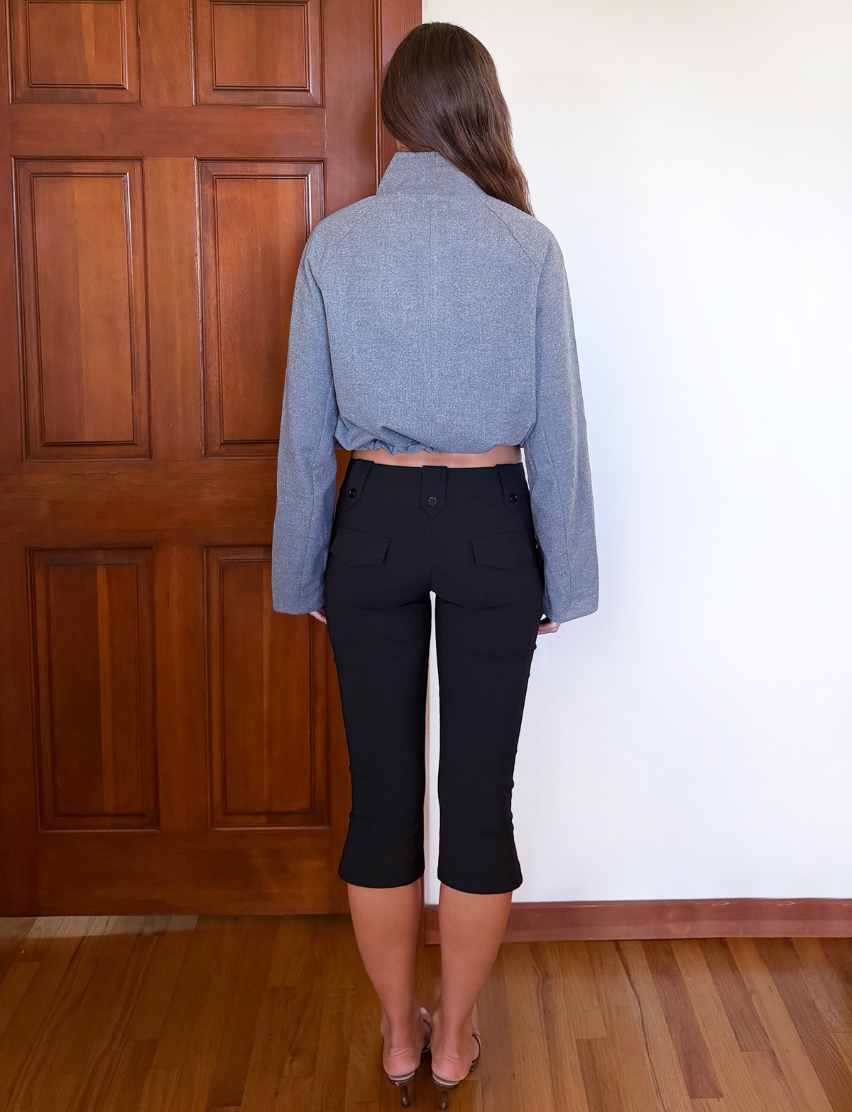 A woman with long brown hair stands indoors on a wooden floor, wearing the TIGERMIST LENNEX JACKET in heather charcoal with a mock neck collar and black cropped pants, facing away from a wood-paneled door and white wall.