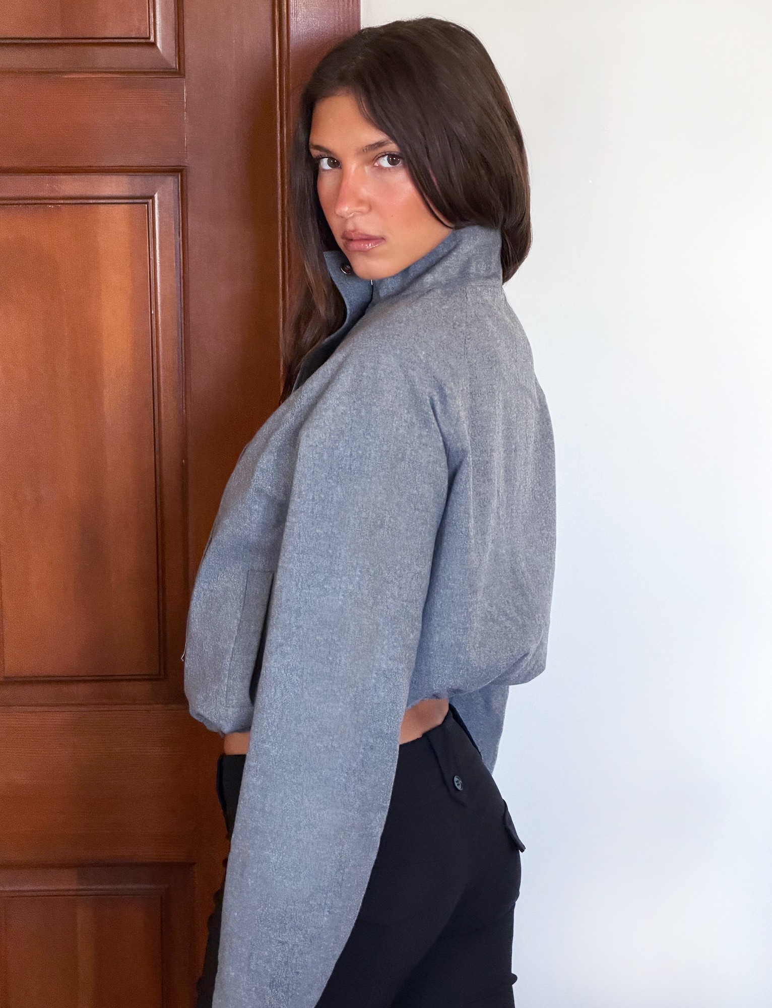 A woman with dark hair glances over her shoulder at the camera, wearing the TIGERMIST LENNEX JACKET in heather charcoal with a mock neck collar, paired with black pants.