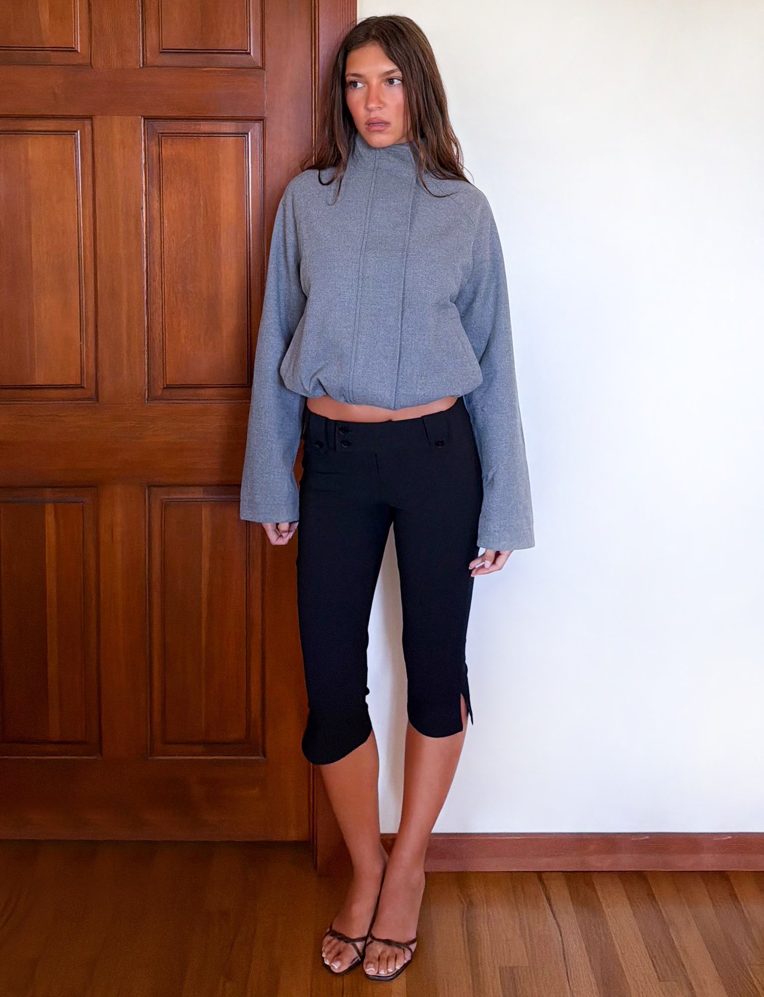 A woman stands indoors by a wooden door, wearing the TIGERMIST LENNEX JACKET in heather charcoal, paired with cropped black pants featuring small side slits and black heeled sandals. She has long brown hair and a neutral expression.