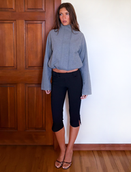 A woman stands indoors by a wooden door, wearing the TIGERMIST LENNEX JACKET in heather charcoal, paired with cropped black pants featuring small side slits and black heeled sandals. She has long brown hair and a neutral expression.