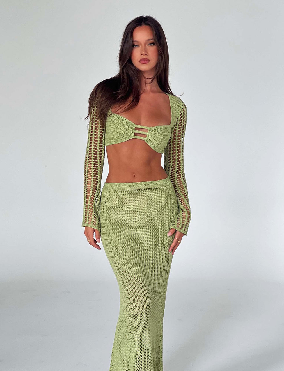 A woman with long, dark hair stands against a plain white background. She is wearing the MACAULEY TOP - GREEN in pastel green from TIGERMIST, a light green knit two-piece outfit featuring a long-sleeve crop top with cutout details and a matching long skirt. She stares directly ahead with a neutral expression.