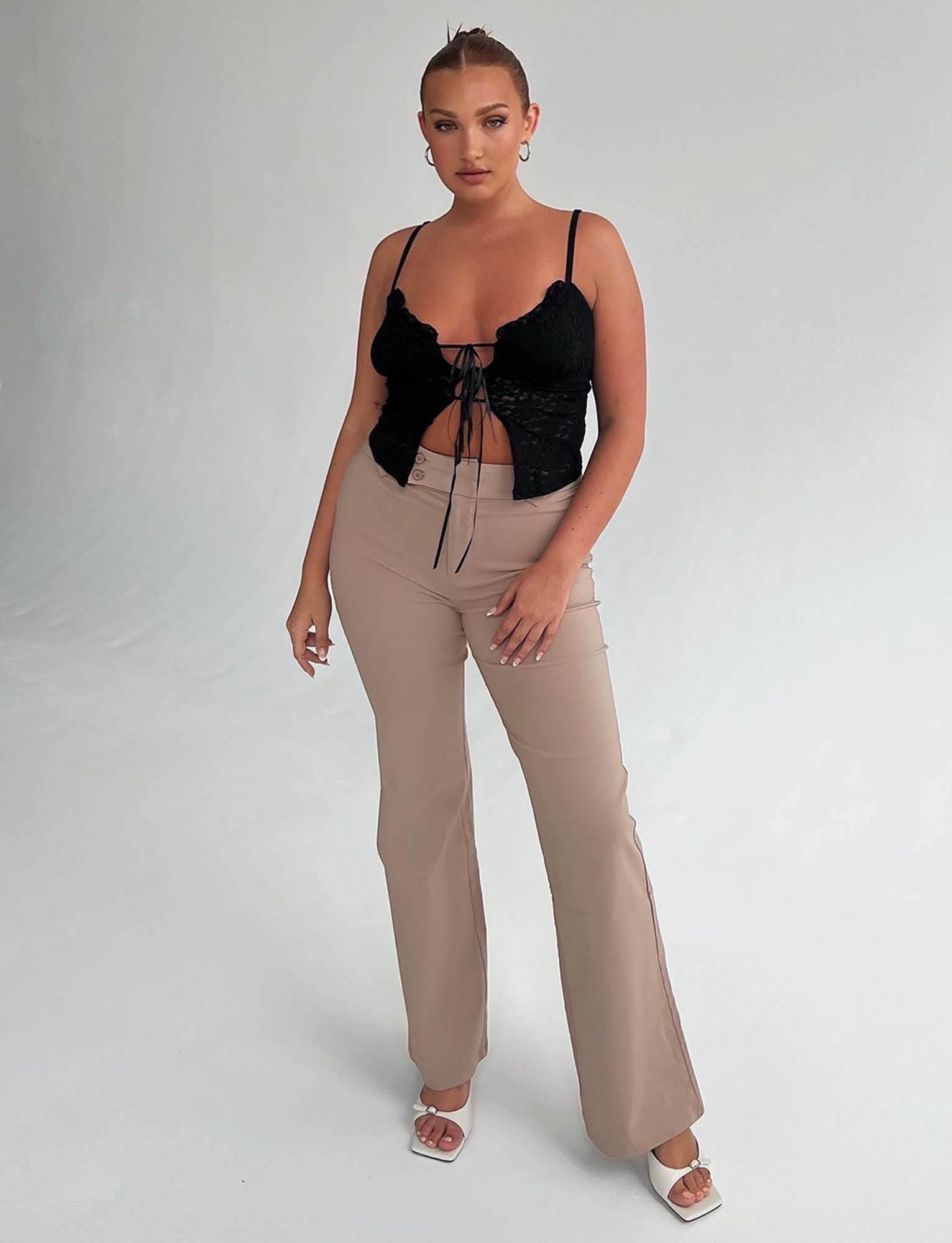 A person with tied-back hair is posing in a studio against a plain background, stylishly dressed in a chic black lace-up crop top from Tiger Mist, paired with the TIGERMIST KITTIE HIGH RISE PANT in beige. They complete the look with white heeled sandals and large hoop earrings, opting for minimal makeup.