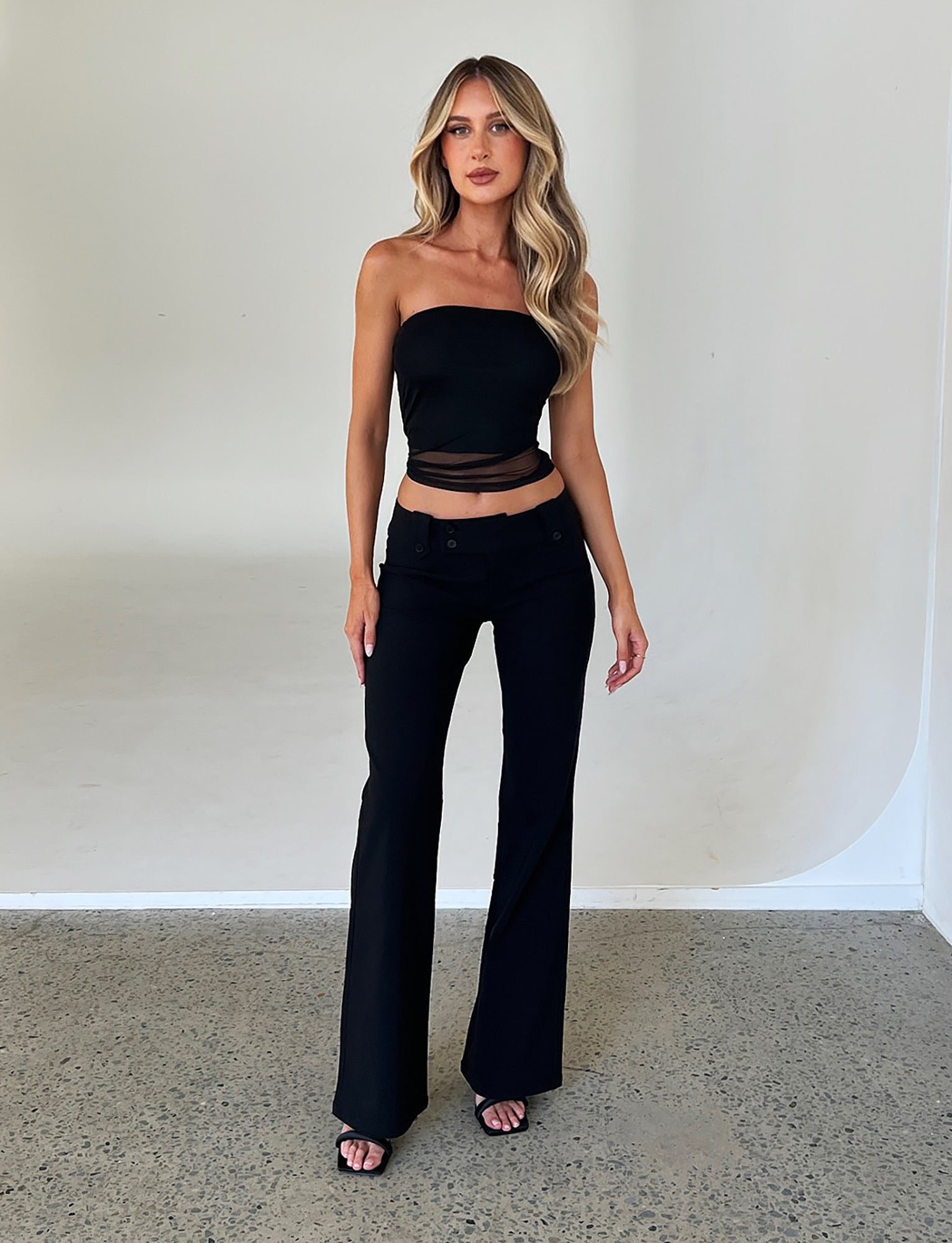 A woman stands indoors against a white backdrop, wearing the HARRIETT TUBE TOP in black and matching black flared pants from TIGERMIST. She has long, wavy blonde hair and wears black open-toe heels. She stands confidently with her arms relaxed at her sides, looking at the camera.