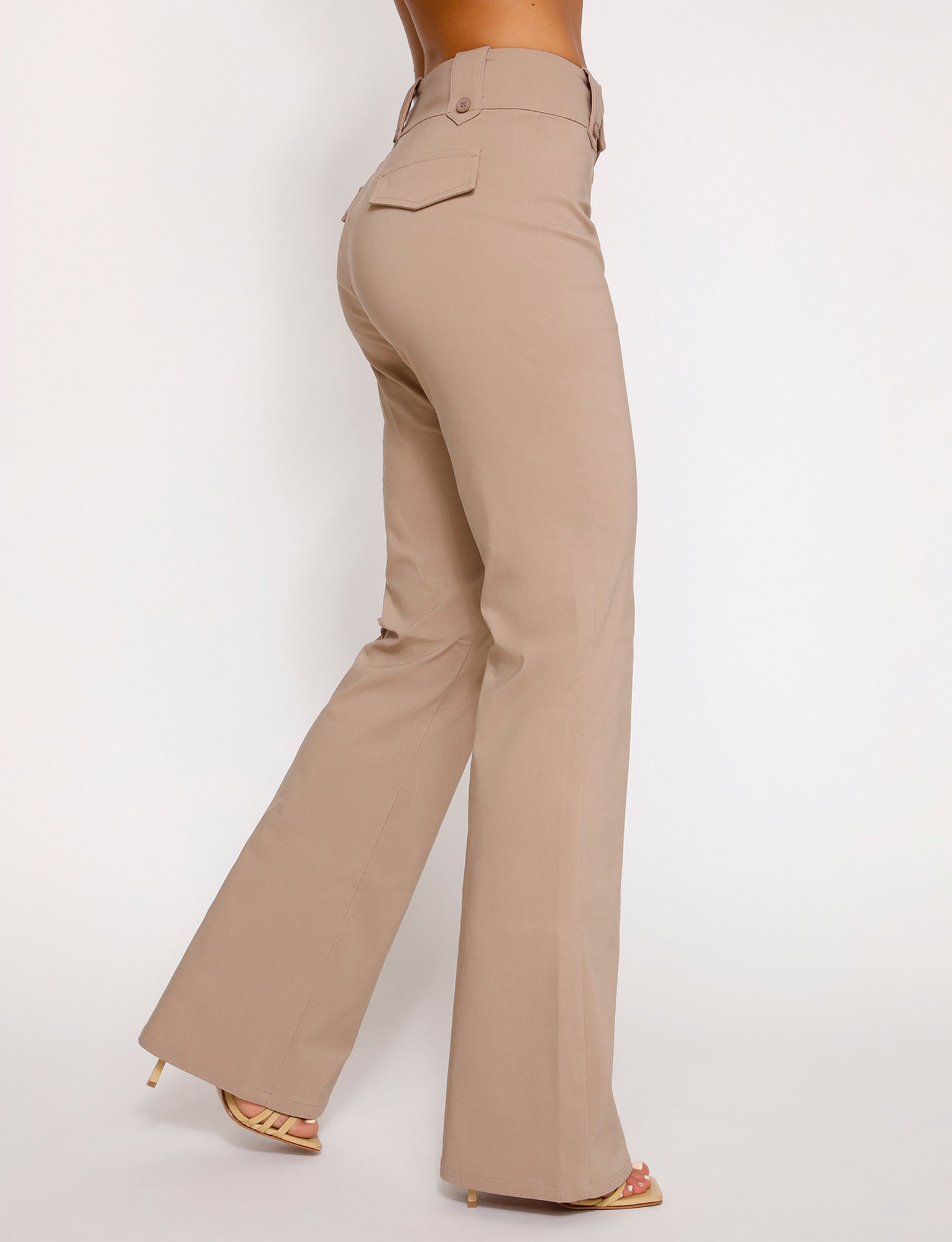 A person wearing the KITTIE HIGH RISE PANT in beige from TIGERMIST and light-colored high-heeled sandals is shown from the waist down against a plain white background. The pants feature a buttoned pocket flap on the back.