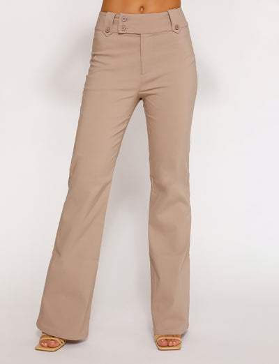 A person is modeling the Kittie High Rise Pant - Beige from TIGERMIST. The high-waisted, flared pants feature a button detail at the waist and offer a smooth, fitted look. The model pairs them with open-toed shoes against a plain white background.