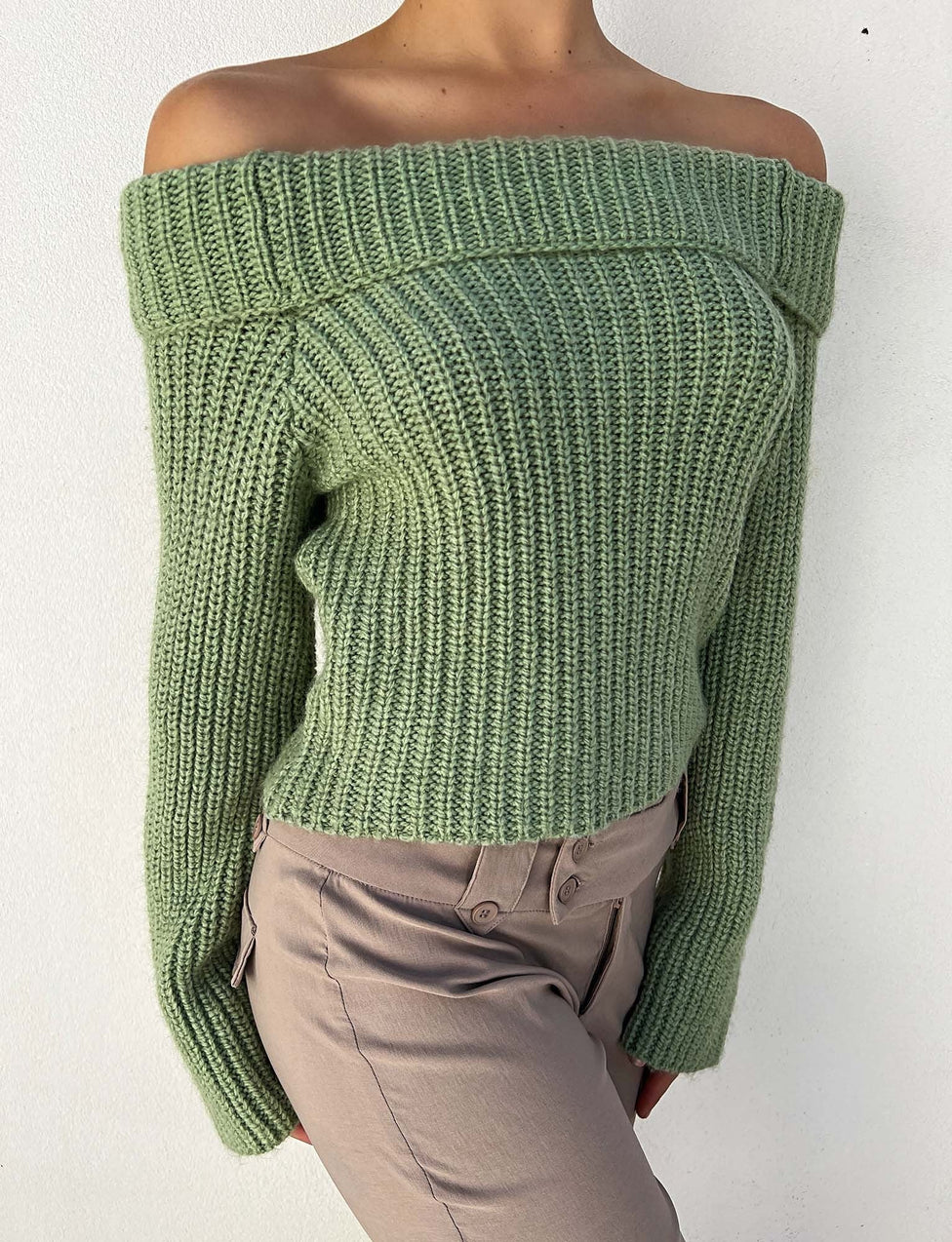 A person is wearing the ASHLYN JUMPER - GREEN from TIGERMIST, paired with beige pants. Only the torso is visible against a plain white background. The off-shoulder, light green, ribbed knit sweater drapes comfortably and has long sleeves.