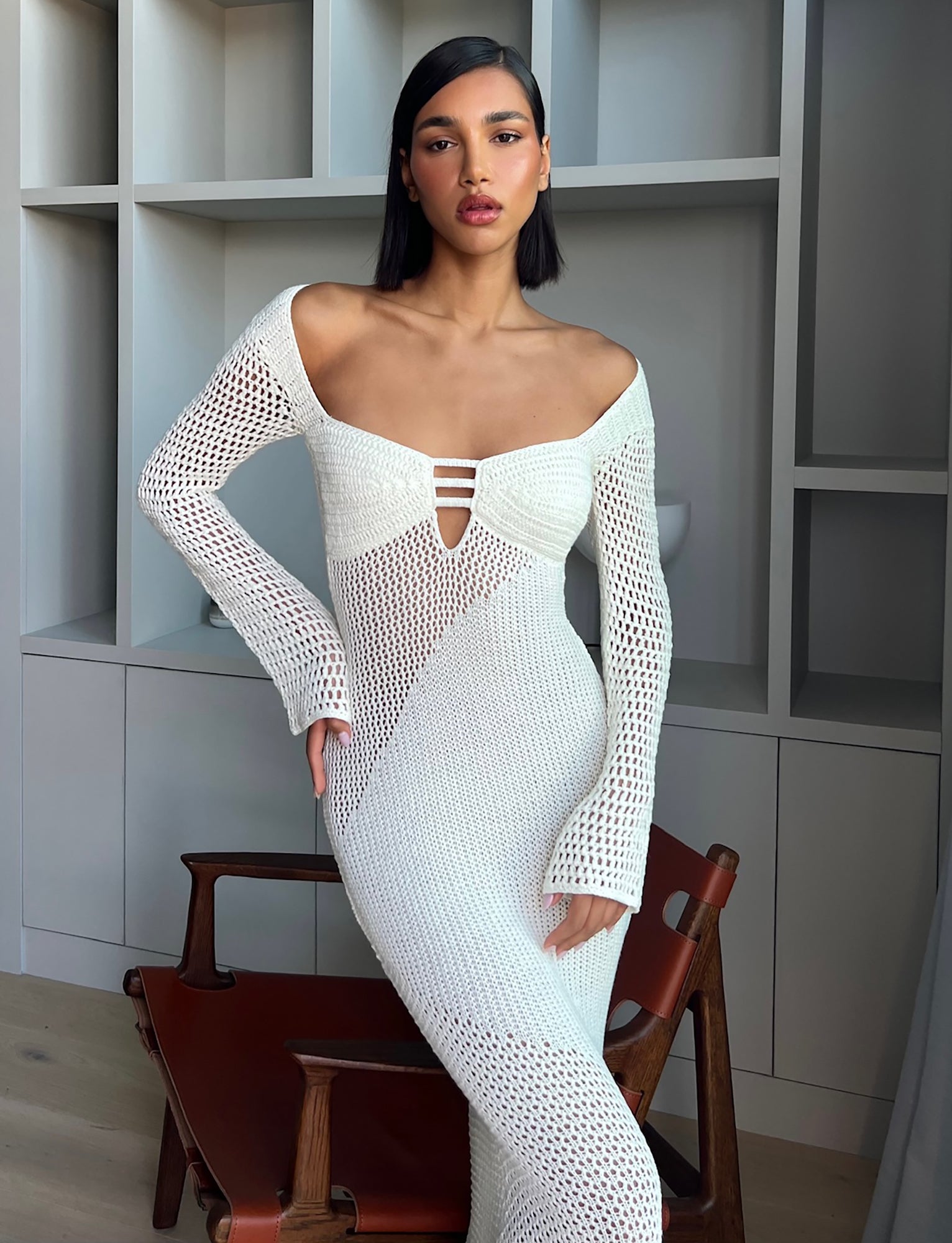 A woman with straight dark hair stands confidently in a room with white shelving. She wears the MACAULEY DRESS - WHITE : CREAM, a long-sleeve, crocheted dress with subtle cutouts by TIGERMIST. Leaning on the back of a wooden chair with her left hand, she gazes directly at the camera.