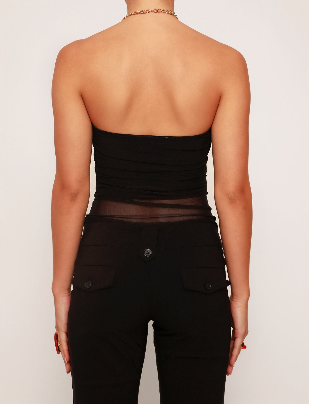 A person is standing with their back facing the camera, wearing a striking TIGERMIST HARRIETT TUBE TOP in black featuring a sheer section in the middle, paired with fitted black pants with buttoned back pockets. Their arms rest by their sides against a plain white background.