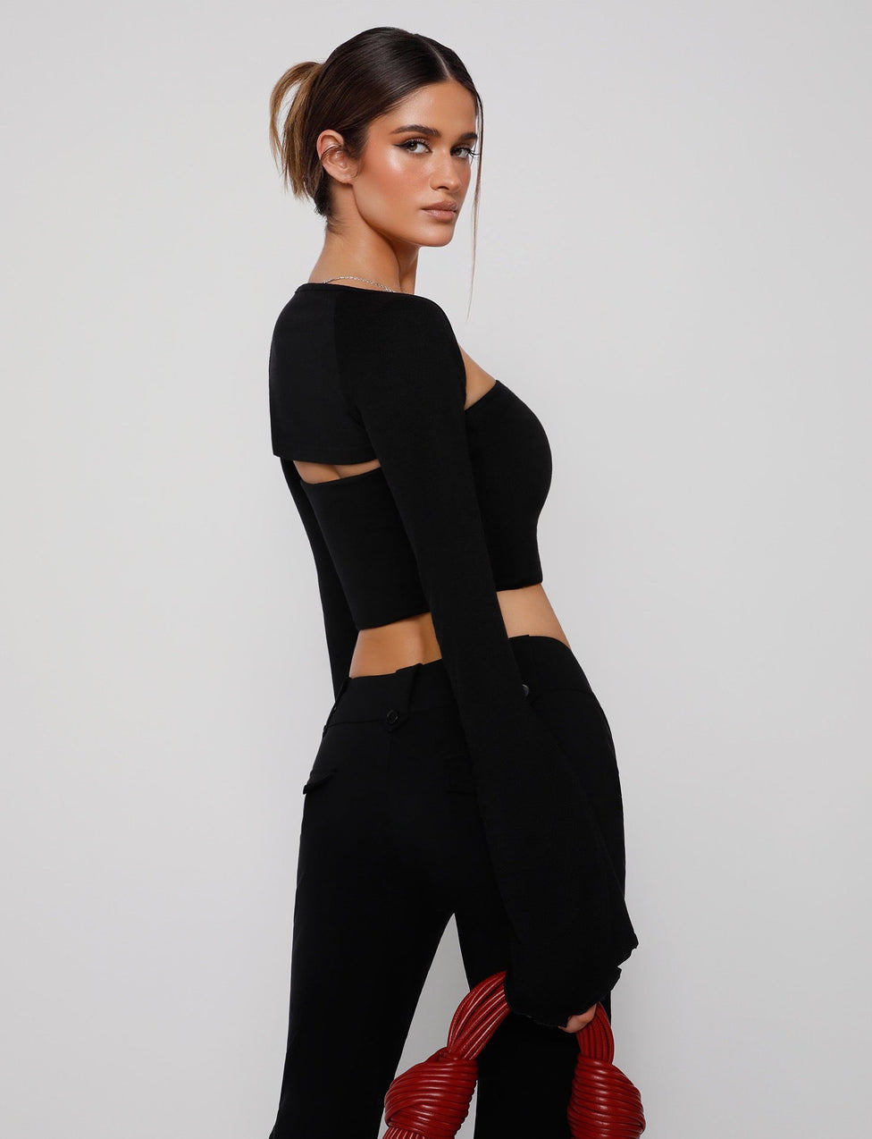 A woman is standing against a plain white background. Her hair is tied back, and she's wearing the TIGERMIST SOPH TWO PIECE TOP - BLACK with a unique cut-out design on the back. Pairing it with black pants, she holds a coiled red accessory in her left hand.