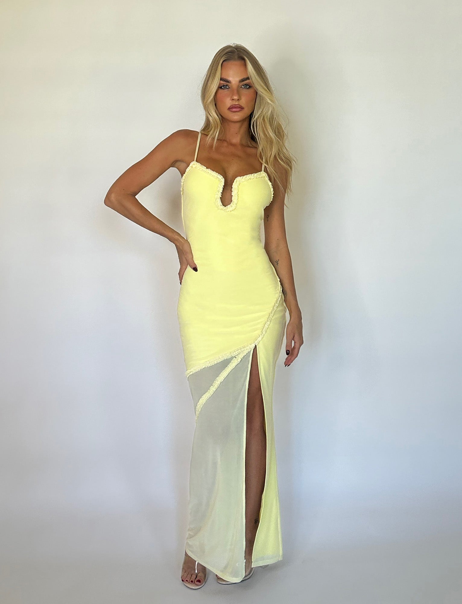 A woman with long blonde hair wears the MIARA DRESS - YELLOW from TIGERMIST, a fitted, pale yellow sleeveless dress featuring a deep neckline and a high slit. She poses against a white background with one hand on her hip and the other arm relaxed by her side, standing in nude high heels.