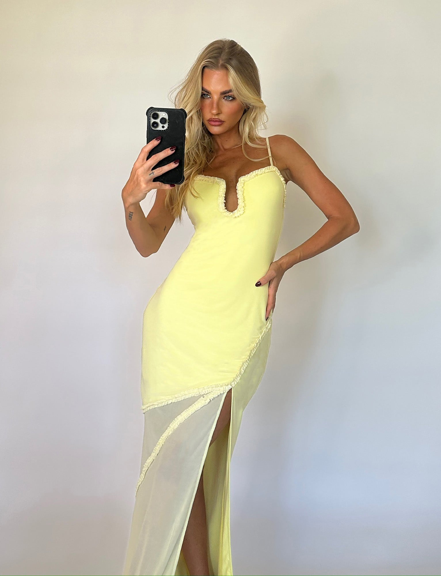 A woman with long blond hair takes a mirror selfie while wearing the pale yellow MIARA DRESS from TIGERMIST, featuring a v-neckline and a high side slit. She stands against a plain white background and holds her phone in one hand.