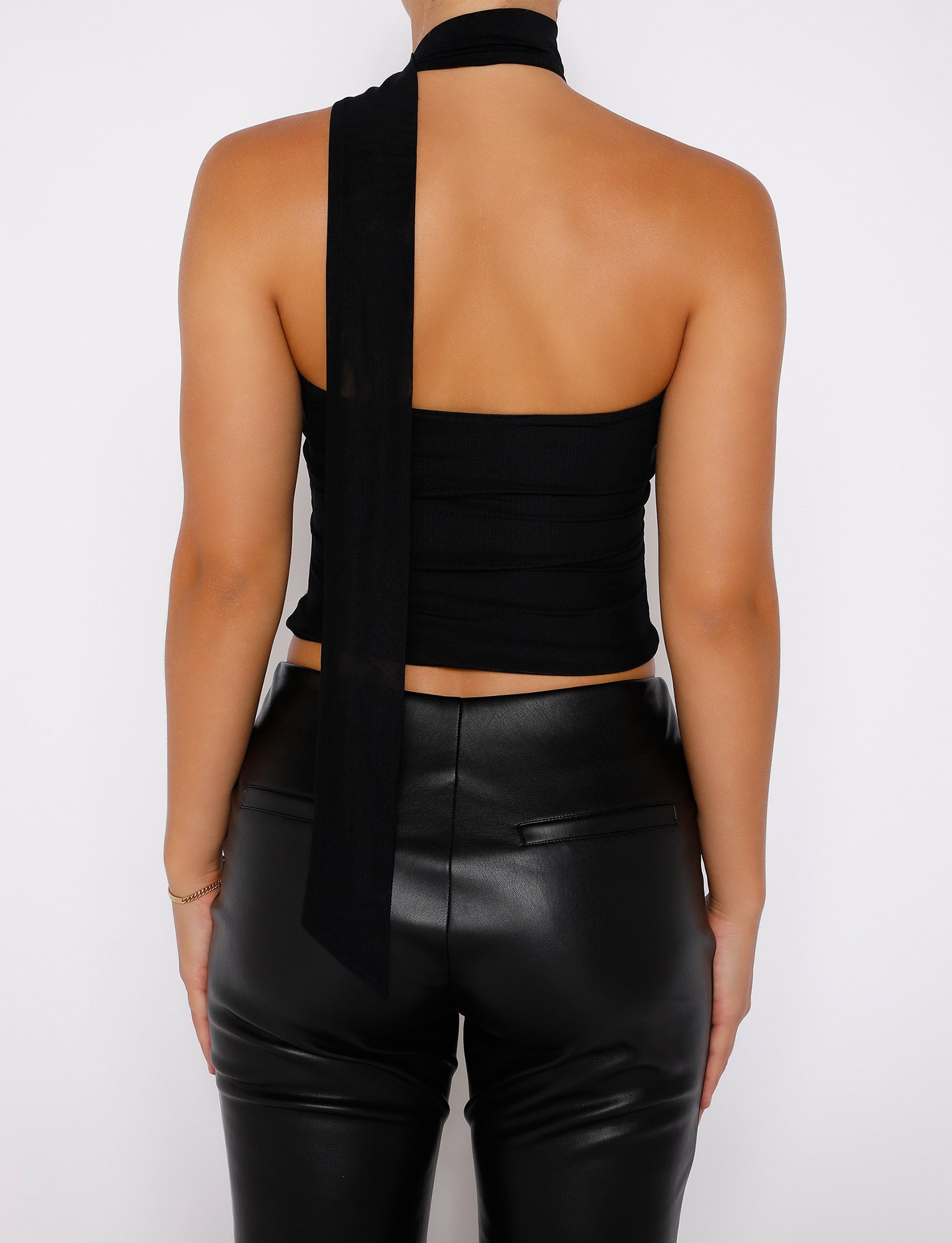 A person with light brown skin is seen from the back, wearing the TIGERMIST CHRYSLER TOP - BLACK, featuring a one-shoulder design with a long scarf-like strap draping down the back, paired with shiny black leather pants. The person's hair is tied up. The background is plain white.