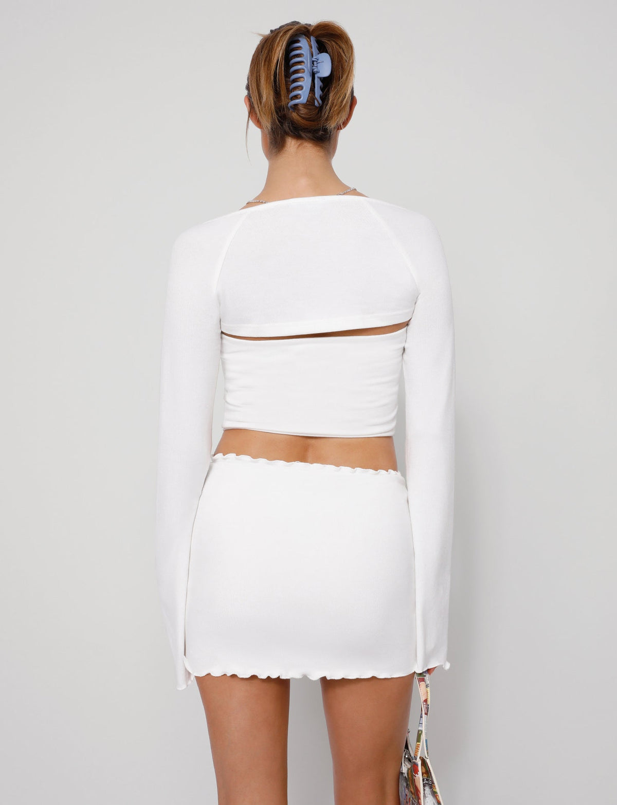 A person facing away from the camera, wearing the chic TIGERMIST SOPH TWO PIECE TOP - WHITE, which comprises a long-sleeve crop top and a matching high-waisted skirt. Their hair is tied back with a blue hair clip, and they are holding a colorful handbag at their side. The background is plain.