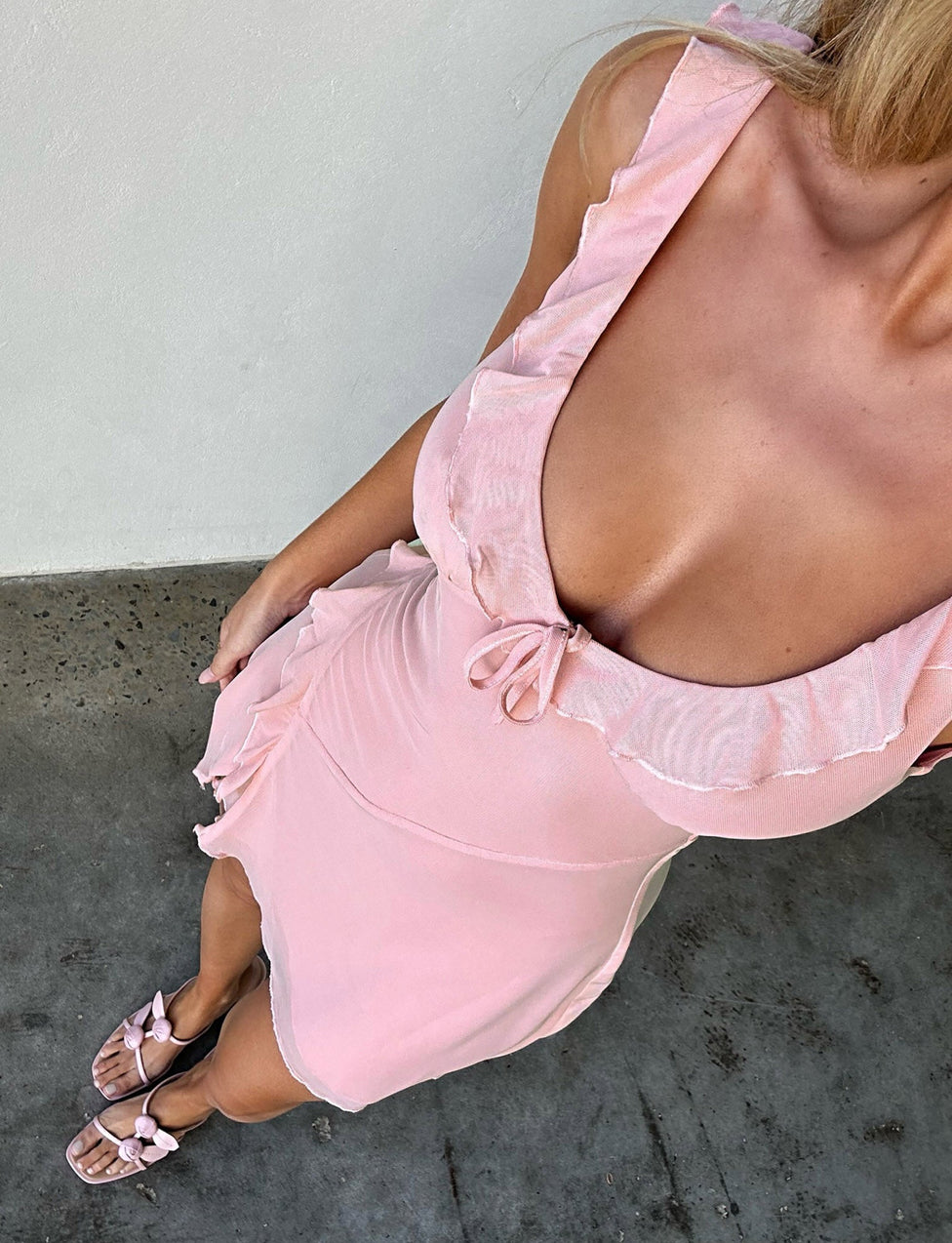 A woman in a dusty pink IMRA DRESS by TIGERMIST, featuring ruffled edges and a plunging neckline, stands on a concrete surface. She pairs her outfit with matching pink sandals adorned with bow accents. The picture is taken from above, highlighting her ensemble and upper body.