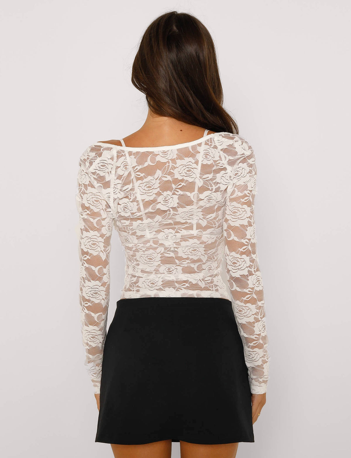 A woman with long brown hair, facing away, is wearing a TIGERMIST CHRISTY LONG SLEEVE TOP in white lace, paired with a short black skirt. The top features a floral pattern and is slightly transparent. She stands against a plain white background.