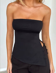 A person wears the elegant, strapless INDIRA TOP - BLACK by TIGERMIST paired with black pants. The top showcases a unique asymmetrical design with side buttons. The image focuses on the upper body and waist, emphasizing the chic and minimalist fashion choice.