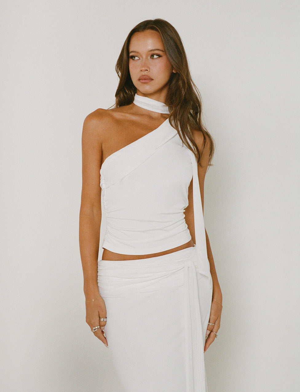 A woman with long, brown hair stands in front of a white background. She is wearing an elegant TIGERMIST ALLIRA TOP - WHITE with a choker-style neck strap. Her hands rest at her sides, and she has a calm, composed expression on her face.