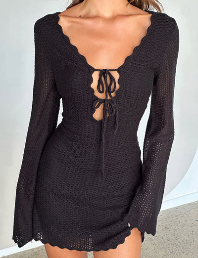 A person wearing the TEREZA DRESS - BLACK from TIGERMIST, featuring a scalloped neckline, long sleeves, and a front cut-out tied with a bow. The background appears to be a simple indoor setting.