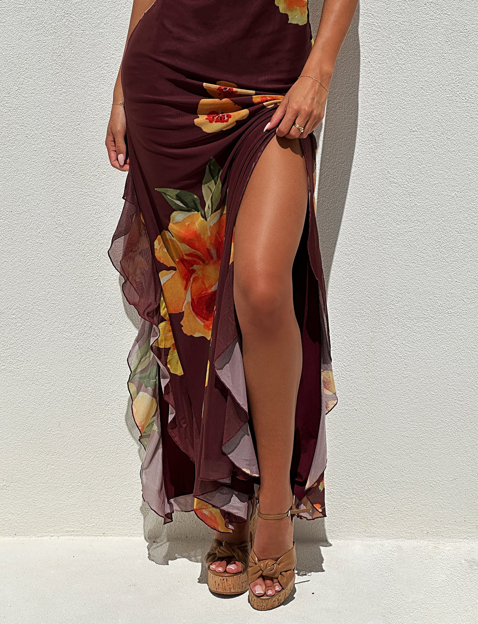 A person wearing the Tigermist Neve Dress - Red: Macro Floral: Burgundy Floral with a high slit and tan platform sandals stands against a white wall. The dress features large yellow and orange flowers and ruffled details. Only the lower half of the person is visible in the image.