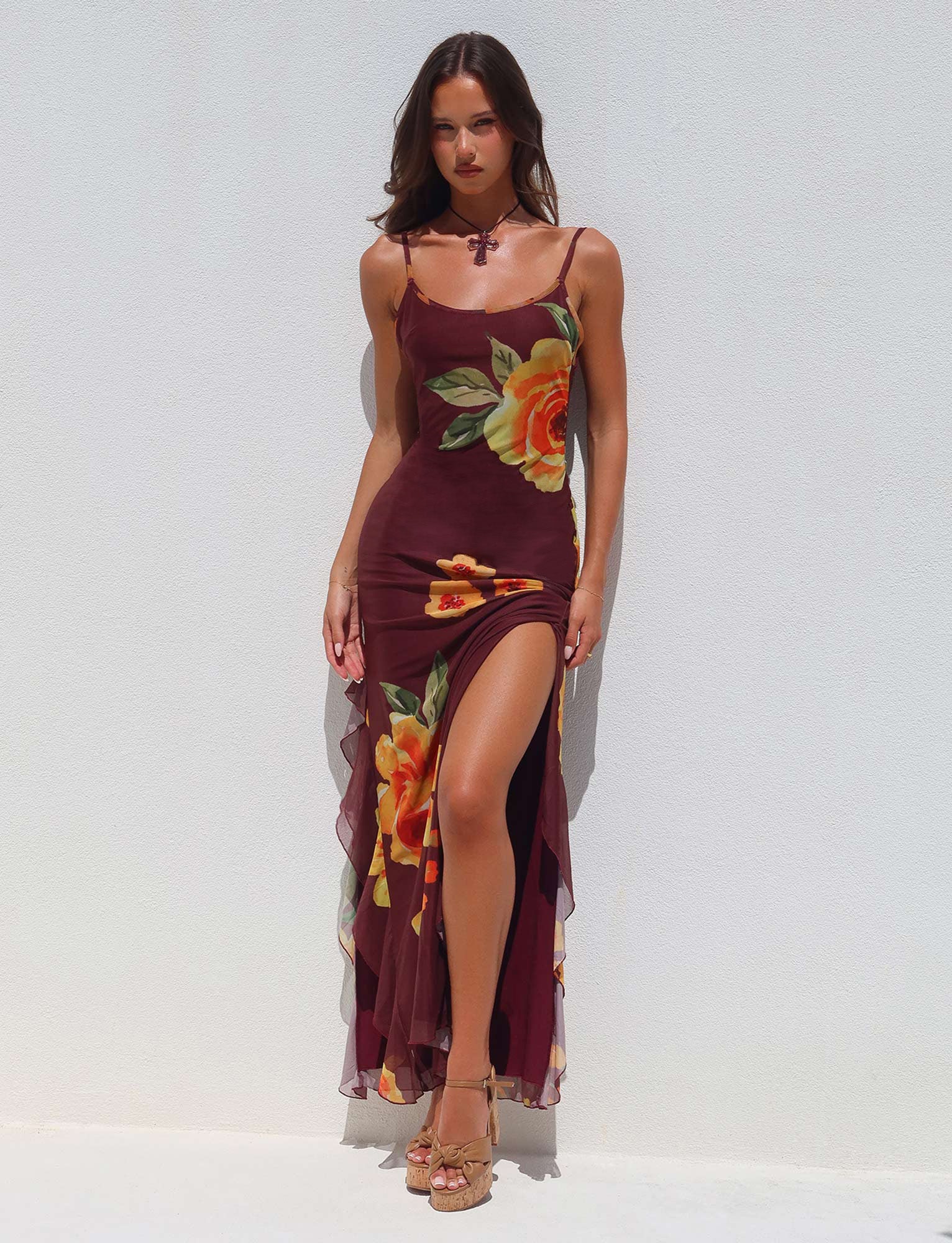 A woman in the NEVE DRESS - RED : MACRO FLORAL : BURGUNDY FLORAL by TIGERMIST stands against a plain white background. The dress features spaghetti straps and a high slit, revealing one leg. She wears high-heeled sandals and gazes directly at the camera.