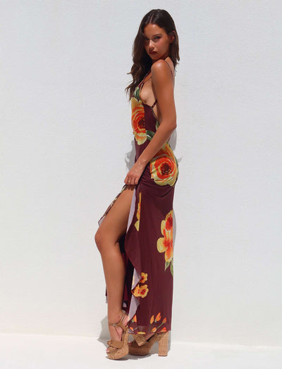 A woman stands sideways against a white backdrop, wearing the NEVE DRESS - RED : MACRO FLORAL : BURGUNDY FLORAL from TIGERMIST. She pairs the outfit with tan wedge sandals and has long brown hair flowing over her shoulders. She gazes directly at the camera with a confident expression.