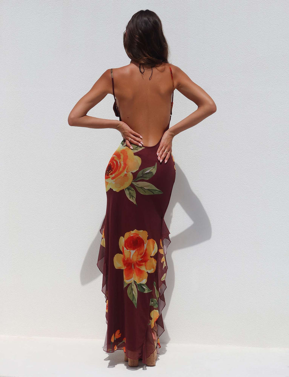 A woman stands against a white wall with her back to the camera, wearing the TIGERMIST NEVE DRESS in red, featuring a macro floral pattern in burgundy. The dress is backless with large orange and yellow flower prints and has thin shoulder straps. She has long hair and is holding her arms slightly bent behind her back.