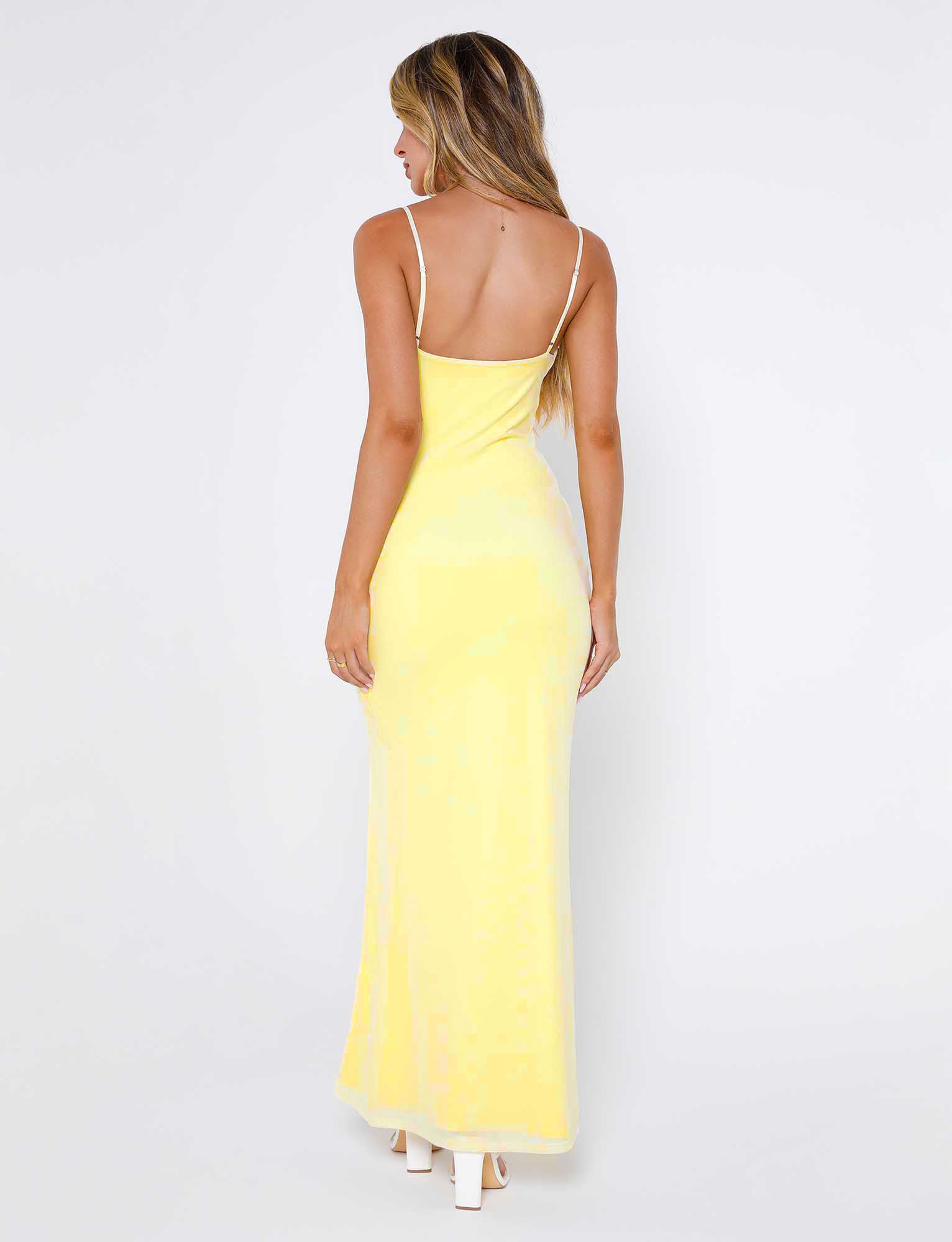 A woman with long, wavy hair stands facing away, wearing the MIARA DRESS - YELLOW : PALE YELLOW : CITRUS from TIGERMIST, a long, sleeveless dress with thin straps. She is also wearing white high-heeled shoes. The background is plain white.
