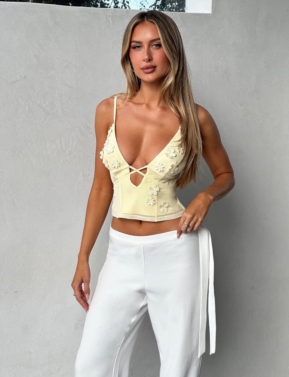 A woman with long blonde hair stands against a gray wall, confidently sporting the KATINA CAMI - YELLOW: BUTTER from TIGERMIST. The light yellow, sleeveless top features flower embellishments and a V-neckline, perfectly paired with her white pants. Her left arm rests on her hip as she gazes assuredly ahead.