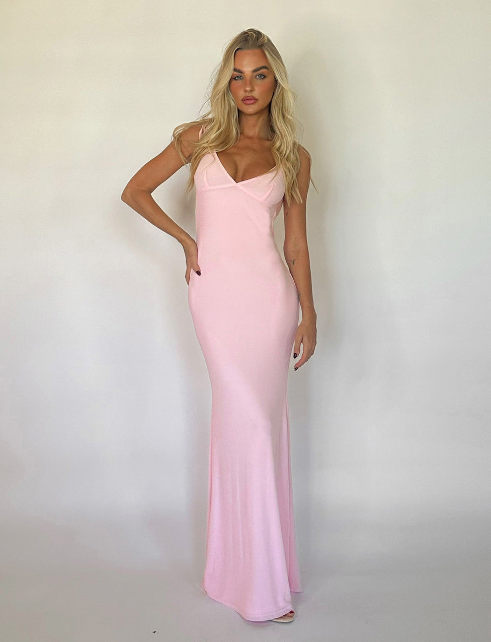 A woman stands against a plain white background, wearing the form-fitting, floor-length KYLAH DRESS in baby pink by TIGERMIST. She has long, wavy blonde hair and a confident expression, with one hand resting on her hip and the other hanging by her side.