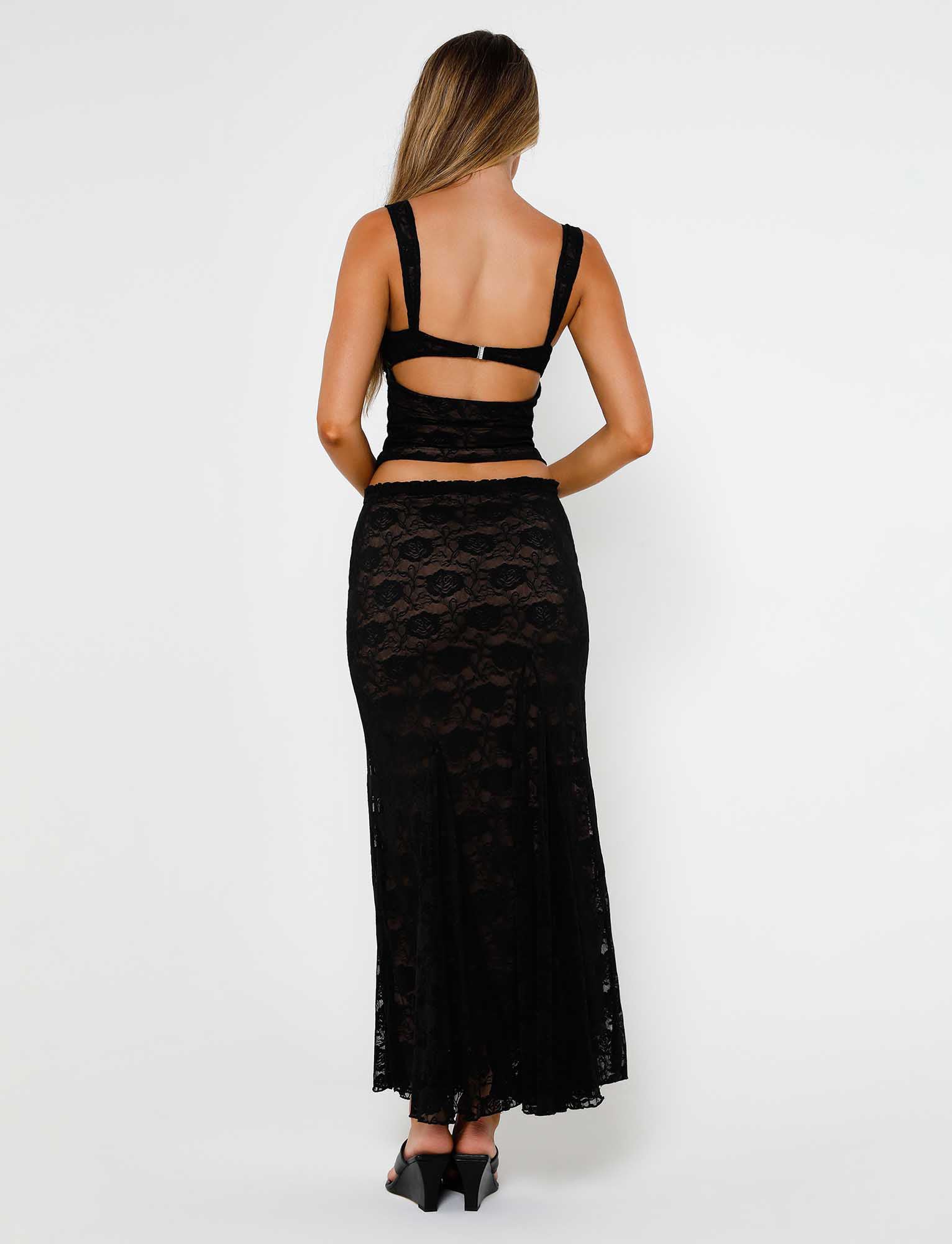 A person with long, light brown hair is turned away from the camera, wearing a black lace top with a strap across the back and the ISADORA SKIRT - BLACK : LACE from TIGERMIST. They are also wearing black heels. The background is plain and white.