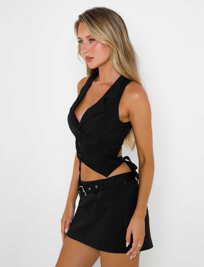 A woman with long blonde hair is wearing a sleek black sleeveless top from TIGERMIST, featuring a collar and buttons, paired with the NADINE SKORT - BLACK. She is posing against a plain white background, looking slightly to the left with a relaxed yet confident demeanor.