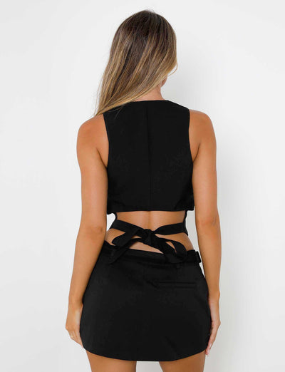 A person with long, light brown hair is facing away from the camera, donning a chic black sleeveless top tied at the back and TIGERMIST's NADINE SKORT in black. The background is plain and white.